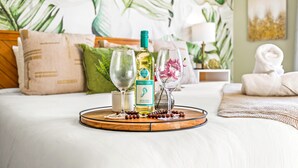 Un vassoio ordinato con una bottiglia di vino, due bicchieri e una piccola ciotola di frutta su un letto con carta da parati a motivi tropicali.