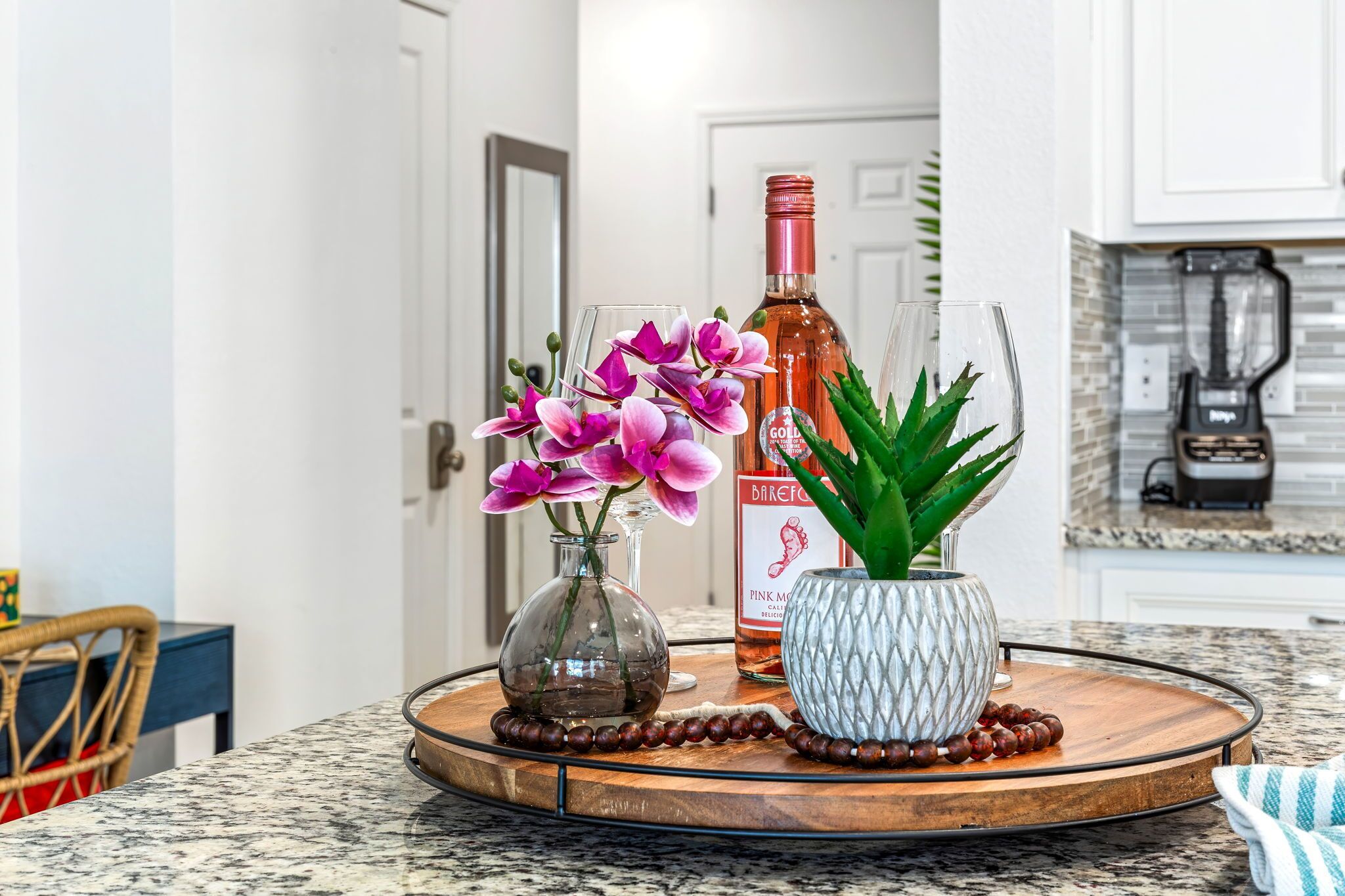 Un comptoir de cuisine sur lequel repose un plateau rond en bois, contenant un vase en verre avec des fleurs violettes, une bouteille de vin rosé et un vase blanc texturé avec des feuilles vertes.