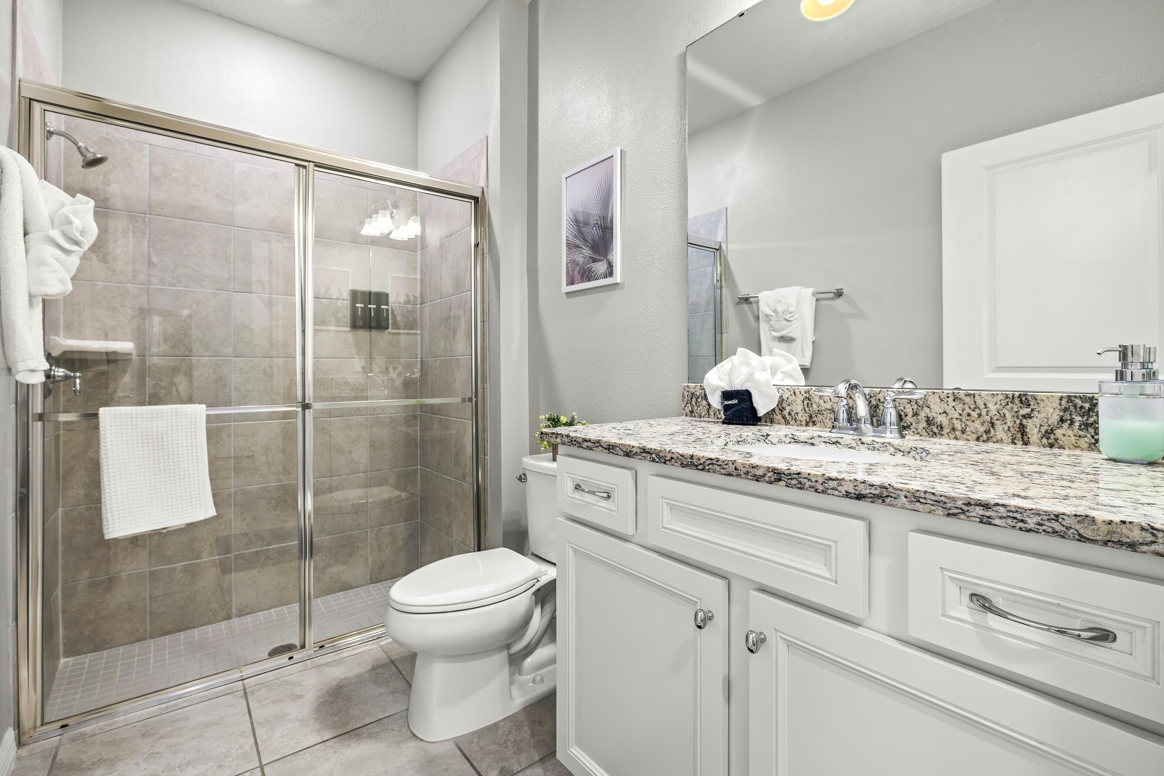 Un baño moderno con cabina de ducha de vidrio, inodoro blanco y encimera de granito con lavabo y espejo.