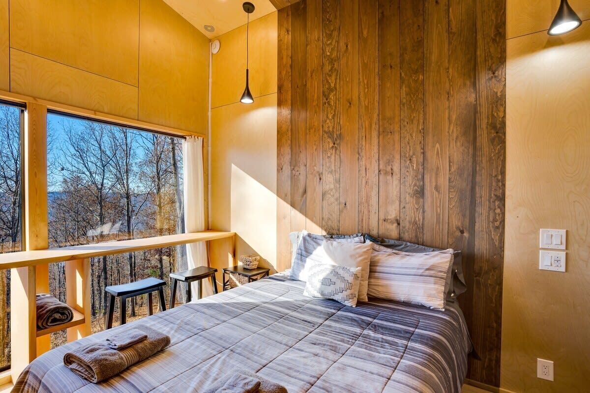 Ein Schlafzimmer mit großem Fenster, Holzwandverkleidung, einem Bett mit gemusterten Textilien und einem kleinen Tisch mit Stuhl.
