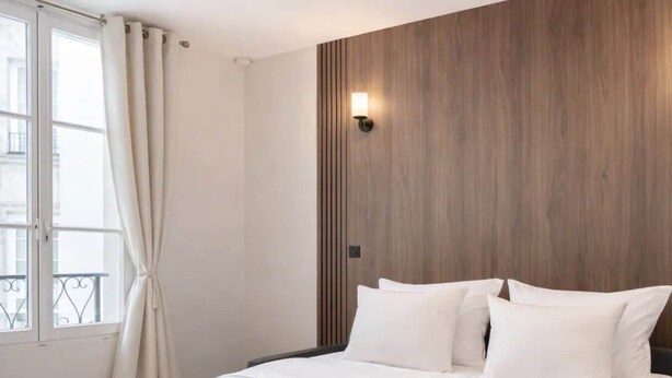 A hotel room with a bed, white linens, a wooden accent wall, a window with curtains, and a wall-mounted light. - Stunning studio with lift - Louvre (Paris)