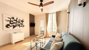 A modern living room with a ceiling fan, a sofa, a coffee table, a TV mounted on the wall, and a window with curtains. - Stunning studio with lift - Louvre (Paris)