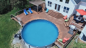 Een rond boven­grond zwembad met een houten terras, omringd door een huis met een groot raam en een overdekt terras.