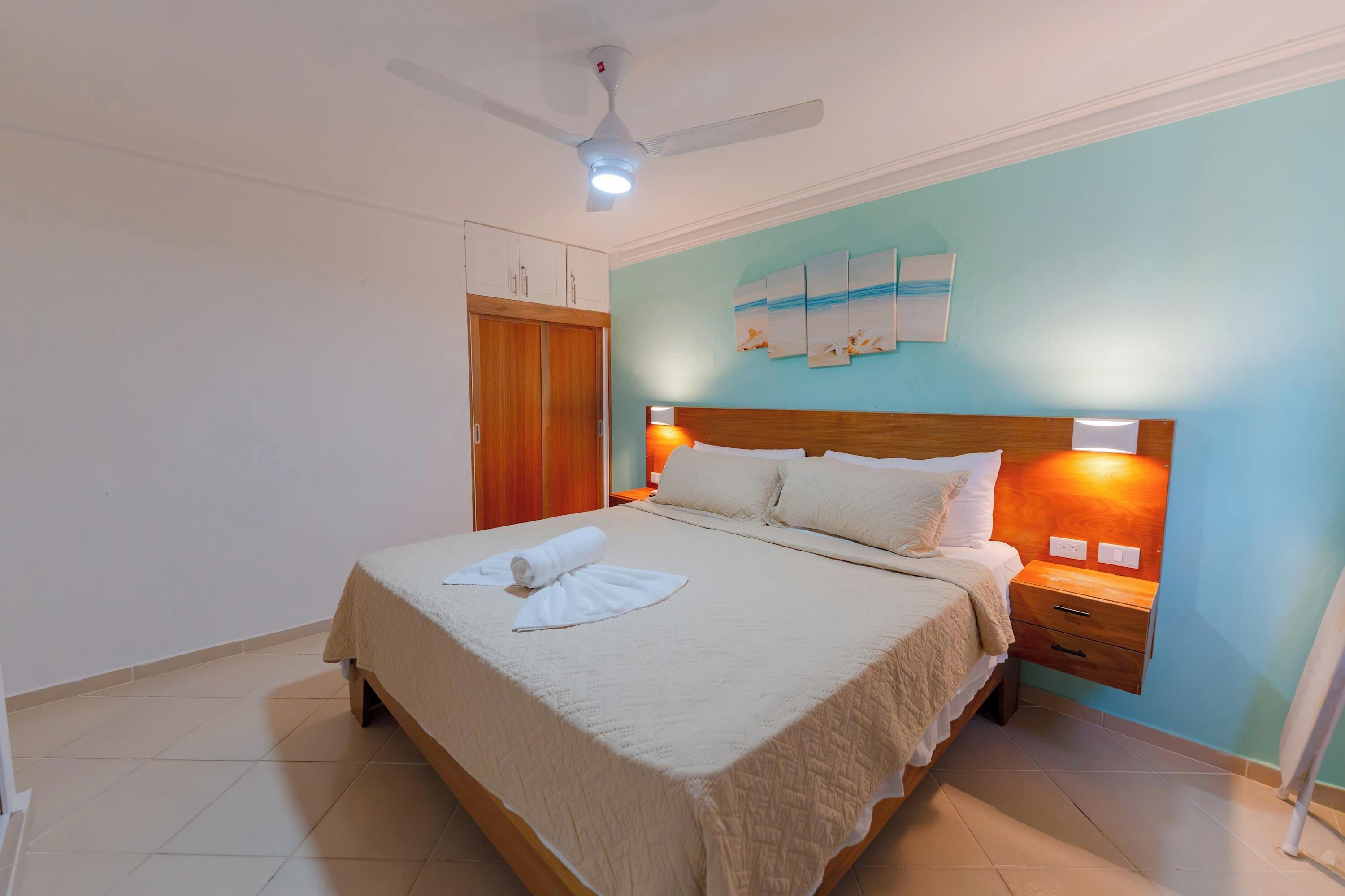 A hotel room with a bed, a nightstand, a ceiling fan, and a wall with artwork.