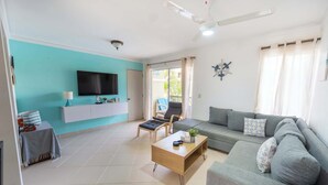 Un salon avec un canapé gris, une table basse en bois, un téléviseur fixé sur un mur turquoise et un ventilateur de plafond.