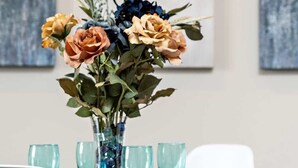 Una mesa de comedor con un florero de flores, cuatro vasos azules y sillas blancas.