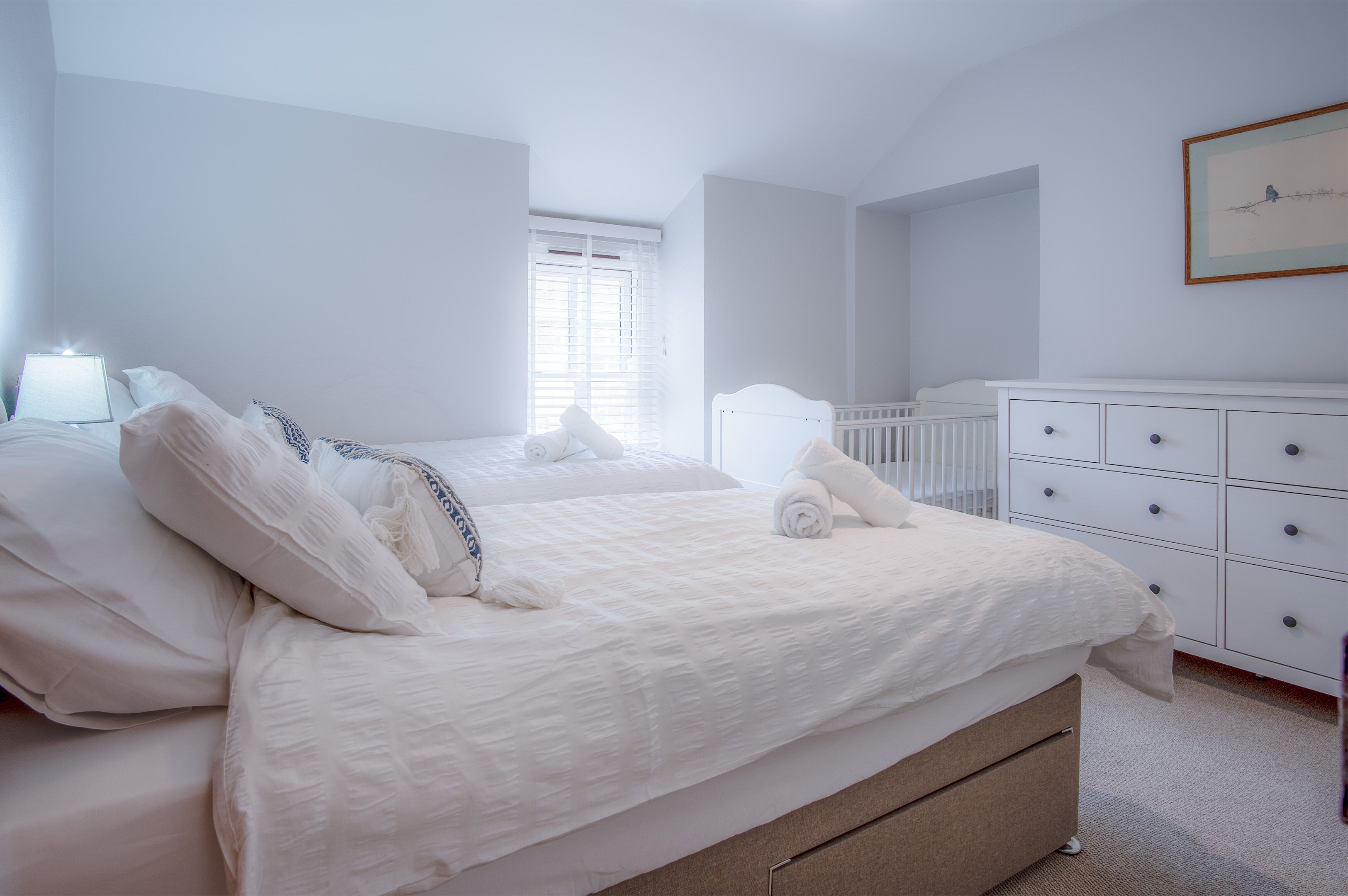 A bedroom with a large bed, a crib, a dresser, and a framed picture on the wall.
