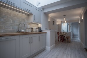 A modern kitchen with white cabinets, a wooden countertop, and a dining area with a table and chairs. - Noe Acre - 3 Bedroom Cottage - Tenby (Tenby)