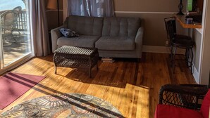 A living room with a grey sofa, a patterned rug, a ceiling fan, and a view through a large window. - Small rustic cabin on KY Lake (sleeps 4) (New Concord)