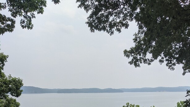 A wooden deck with a table and chairs overlooking a calm lake, surrounded by trees. - Small rustic cabin on KY Lake (sleeps 4) (New Concord)