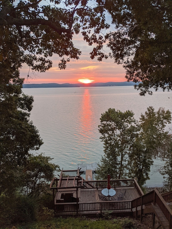 A wooden deck with a table and chairs overlooking a calm body of water during sunset.