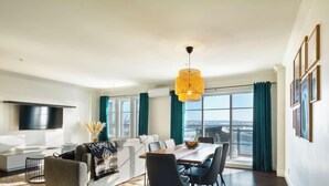 Una sala de estar moderna con televisor de pantalla plana, un comedor con mesa y sillas, y ventanas amplias con cortinas.