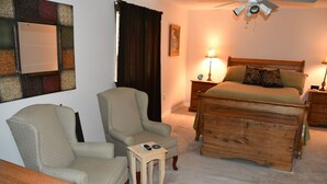 A bedroom with a bed, two armchairs, a wooden dresser, a nightstand, a lamp, and a ceiling fan.