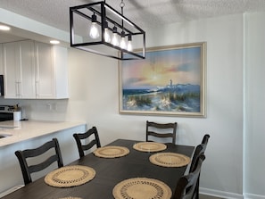Un coin repas avec une table en bois foncé, quatre chaises et des sets de table en fibres tressées. Un tableau représentant un phare sur une plage est accroché au mur.