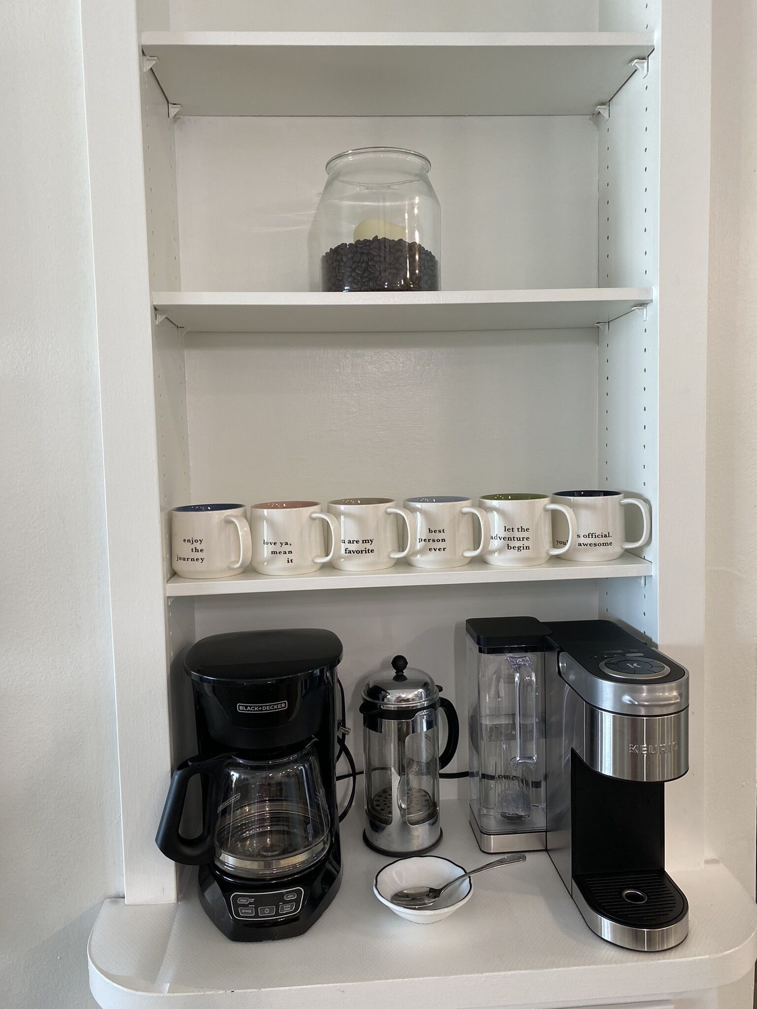 A kitchen shelf with a coffee maker, a water dispenser, and a jar of coffee beans.