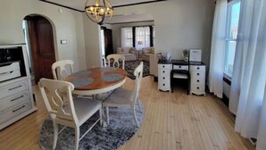 A spacious living room with a round wooden table, white chairs, a chandelier, and a large window with curtains. - SPACIOUS BUNGALOW- 20min to WI Dells/Devils Lake (Portage)