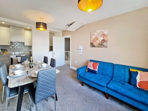 A modern living room with a blue sofa, dining table set for four, and a kitchen in the background.