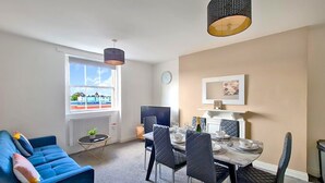 A modern living room with a blue sofa, a dining table with chairs, a TV, and a large window with a view of buildings. - Margaret's Loft by Fortuna Property Portfolio, The Margaret's Loft Apartment (Cheltenham)