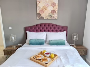 A neatly made bed with a wooden tray of breakfast items, two white pillows, and a purple headboard.