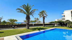 A swimming pool with palm trees, a grassy area, and a view of the sea and buildings in the background.