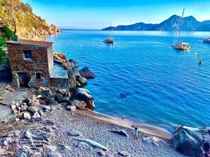 Un paysage côtier avec un bâtiment en pierre, une côte rocheuse, une mer calme et des voiliers.