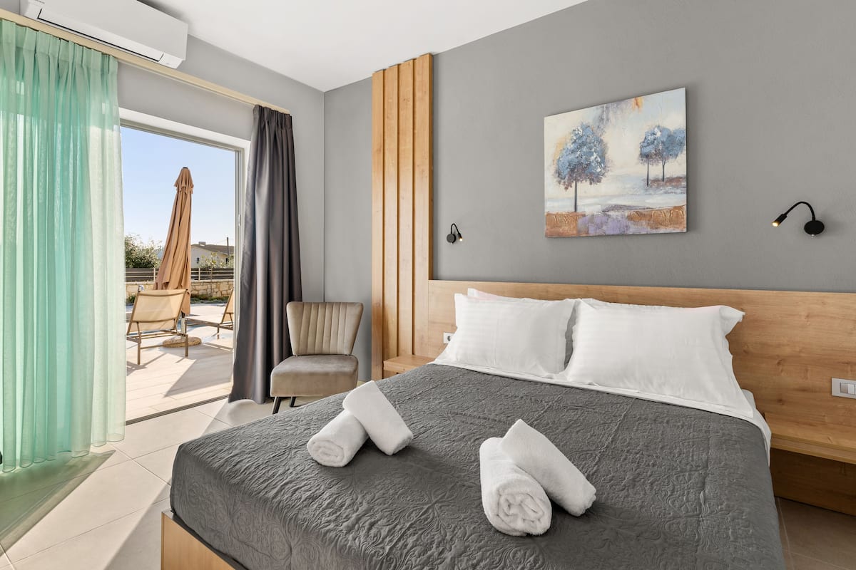 Une chambre d’hôtel moderne avec un grand lit, un tableau représentant des arbres, un fauteuil et une vue sur l’extérieur.