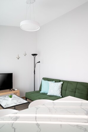 Een moderne slaapkamer met een groene bank, een wit bed, een marmeren bijzettafeltje, een vloerlamp en een televisie op een houten dressoir.
