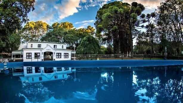 Un edificio blanco y grande con su reflejo en una piscina, rodeado de árboles y un cielo despejado.