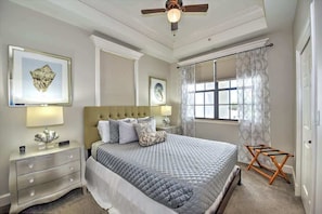 A bedroom with a large bed, a nightstand with a lamp, a ceiling fan, and a window with curtains.