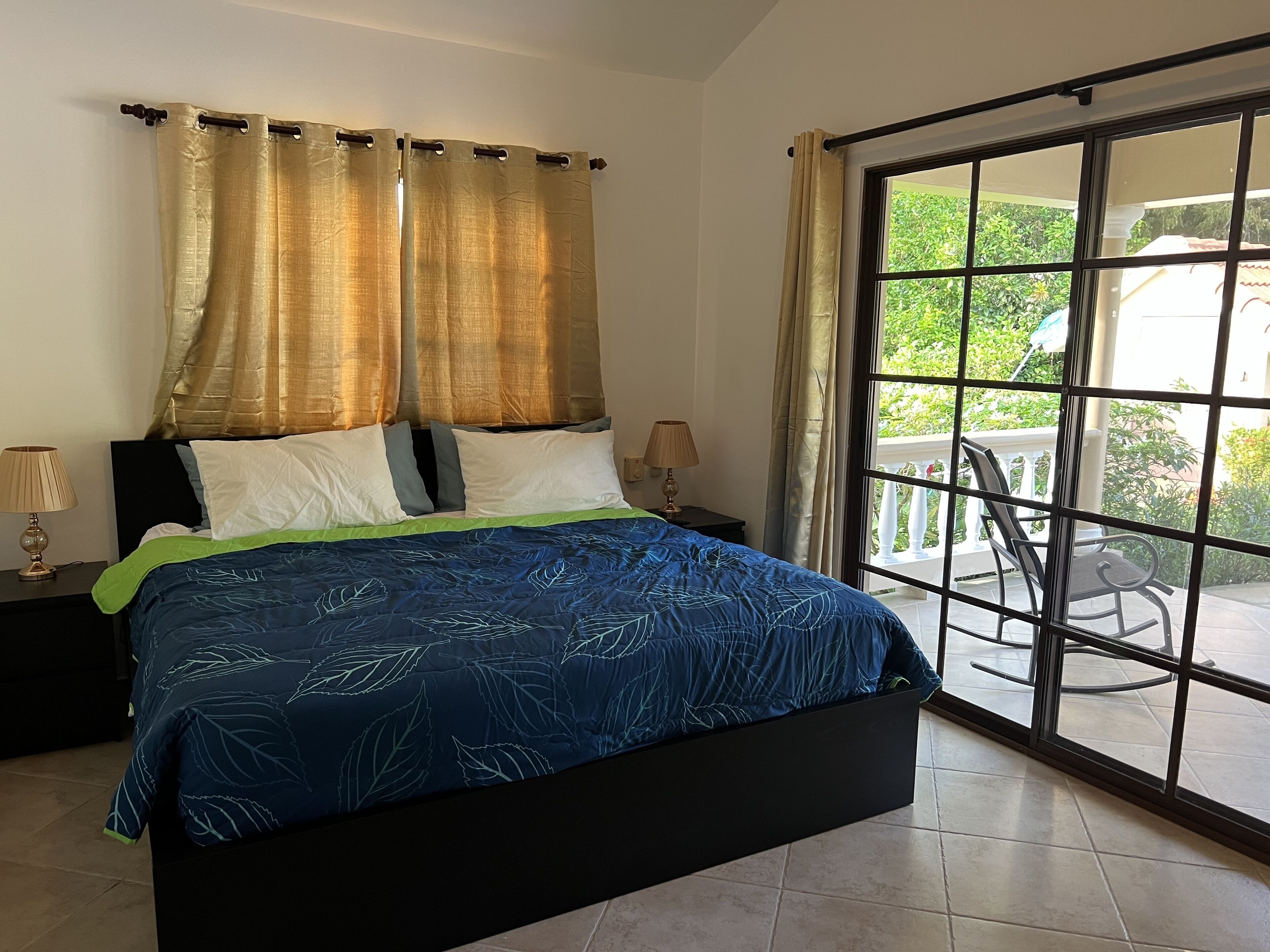 A bedroom with a bed, two bedside tables, a window with a view of greenery, and a balcony with a chair.