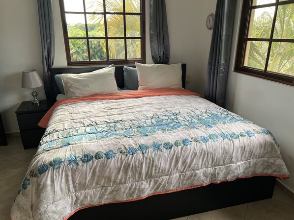 A bedroom with a bed, nightstand, lamp, and a window with curtains. - Charming Hispaniola Villa 23B (Sosúa)