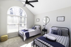 A bedroom with two beds, a ceiling fan, a window with blinds, a bedside table with a lamp, and a small round mirror.