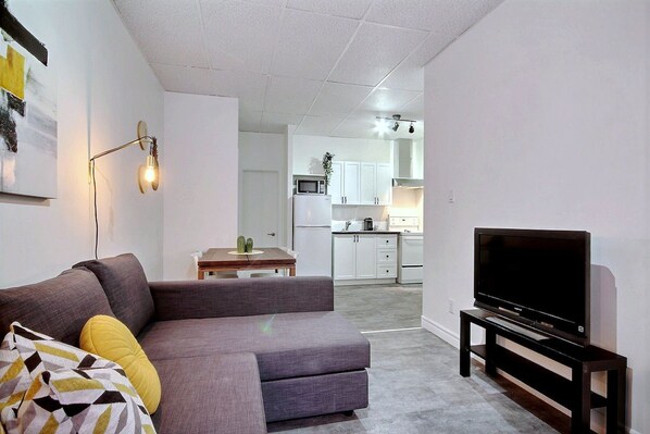A living room with a gray sofa, a television on a stand, and a kitchen in the background.