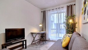A living room with a flat-screen TV on a stand, a small table with a lamp, a sofa with cushions, and a potted plant. - A charming little apartment in the lower town (Québec)