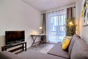 A living room with a flat-screen TV on a stand, a small table with a lamp, a sofa with cushions, and a potted plant. - A charming little apartment in the lower town (Québec)