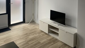 En moderne stue med et fladskærms-tv på et hvidt stativ, en høj gulvlampe og en skydedør til terrassen.