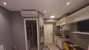A compact hotel room with a bed, a desk, a chair, a small table, a microwave, a TV, and an air conditioning unit. - 123 Quarto - Sé (São Paulo)