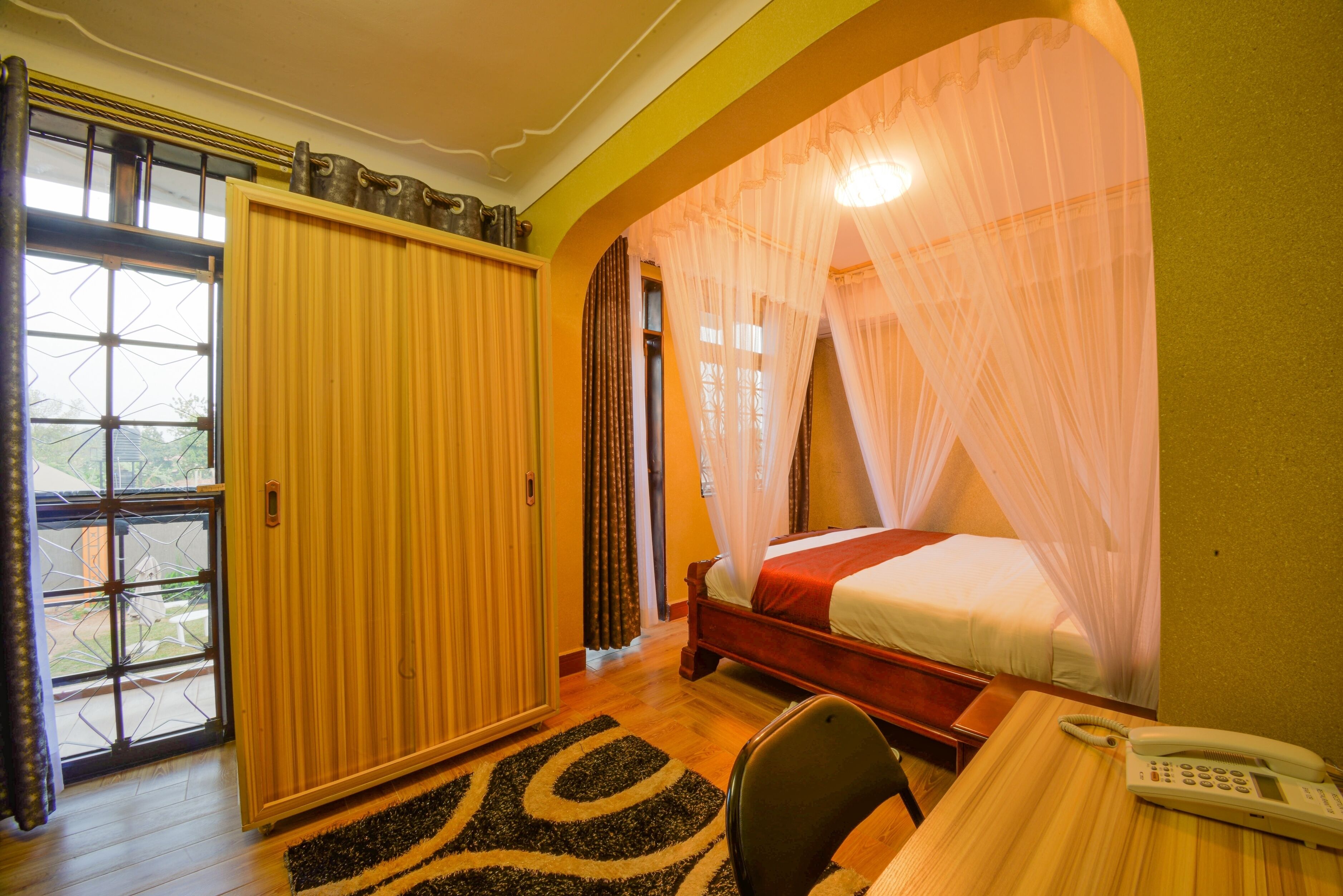 Un dormitorio con una cama bajo una mosquitera, un armario de madera, un escritorio con teléfono y una ventana con cortinas.