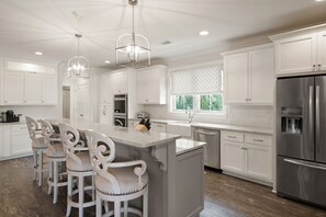 A modern kitchen with white cabinetry, a large island with bar stools, stainless steel appliances, and pendant lighting. - Walk'n on Sunshine - 19 Hendrix Walk (St Simons Island)