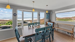 Un espace repas avec une table en bois, des chaises de couleur turquoise et des luminaires suspendus. De grandes fenêtres offrent une vue sur l’océan.