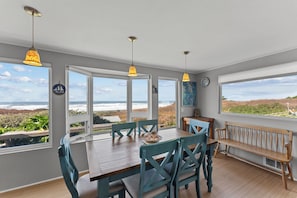 A dining area with a wooden table, teal chairs, and pendant lights. Large windows offer ocean views. - Beach House @ Spirit Cove- Beach Path & SPA (Gold Beach)