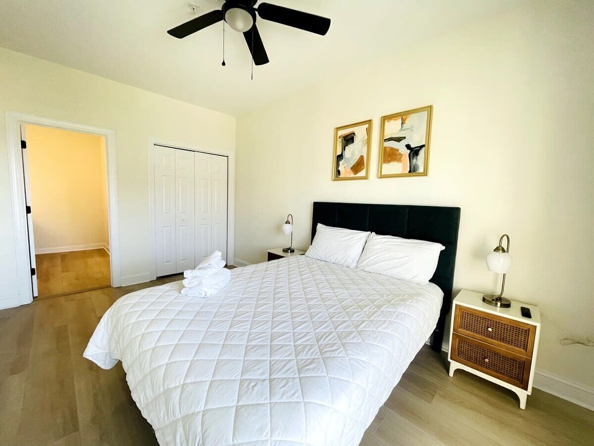 A bedroom with a large bed, a ceiling fan, a nightstand with a lamp, and two framed artworks on the wall.