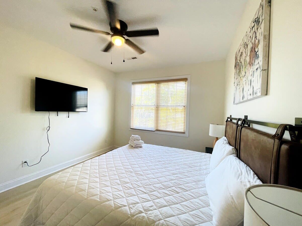 A bedroom with a bed, a ceiling fan, a TV mounted on the wall, a window with blinds, and a painting on the wall.