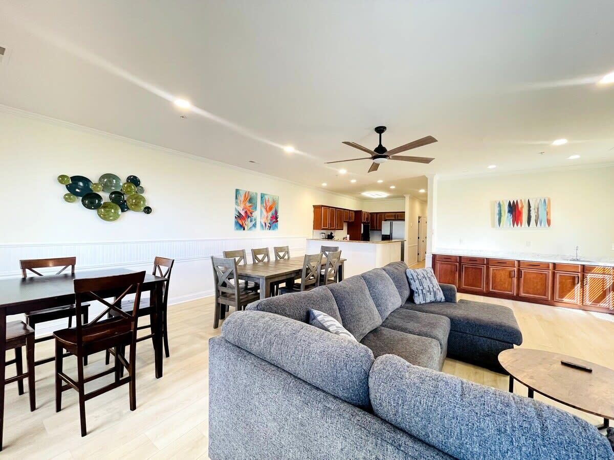 A spacious living area with a large gray sectional sofa, wooden dining table with chairs, and a kitchen in the background.