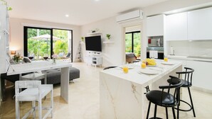 A modern kitchen with white cabinets, a central island with bar stools, a flat-screen TV, and a dining area with a sofa and chairs. - Private Garden Condo by Concierge4u (Playa del Carmen)
