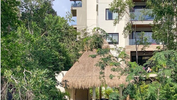 A pool area with a thatched-roof structure, surrounded by lush greenery and a multi-story building in the background. - Private Garden Condo by Concierge4u (Playa del Carmen)