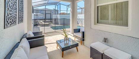 A sunlit patio area with wicker furniture, a glass table, and a view of a swimming pool and houses in the background.