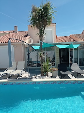 Une piscine avec des chaises longues et un palmier, entourée d’une maison dont le toit est recouvert de tuiles.