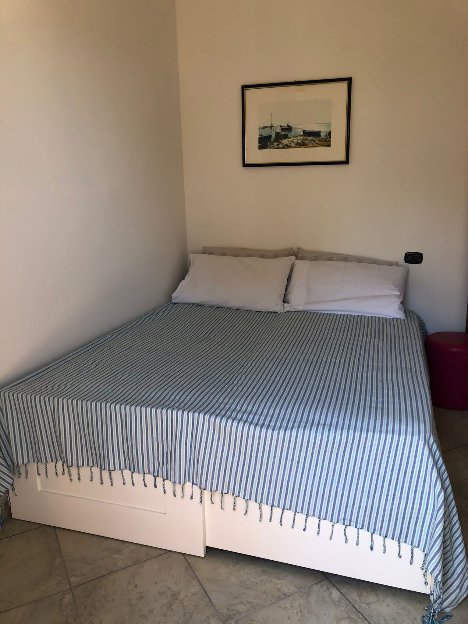 Una cama individual con una colcha a rayas, un cuadro enmarcado en la pared y un taburete rojo.