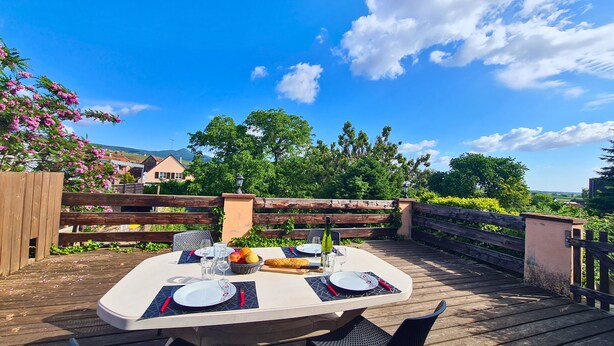 An outdoor dining area with a table set for two, surrounded by a wooden deck and a wooden fence. - Erable – Large Holiday Cottage – Terrace & Parking | Near Colmar and Kaysersberg (Beblenheim)